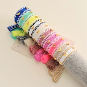 Peut inclure: Une pile de bracelets colorés avec des breloques en forme de papillon dorées et des pompons. Les bracelets sont en tissu tissé et ont un motif rayé.