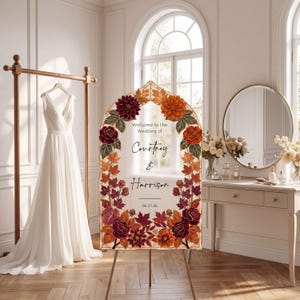 Peut inclure: Panneau de bienvenue de mariage avec des décorations florales. Le panneau affiche les noms "Courtney & Harrison" et la date "06.27.26". Il est orné de fleurs et de feuilles bordeaux, orange et vertes. Une robe de mariée blanche est suspendue à un portant doré.