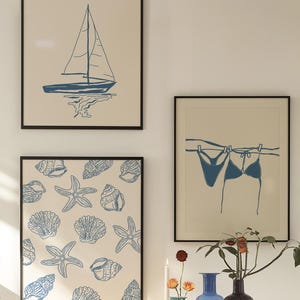 May include: Three framed art prints with a nautical theme. One print features a blue sailboat, another showcases blue seashells and starfish, and the third depicts a blue bikini hanging on a clothesline. All prints have black frames.