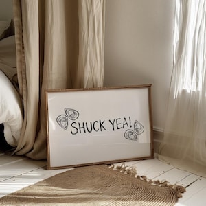 May include: A framed white print with the text "SHUCK YEA!" and oyster illustrations. The frame is a light brown color. The print is leaning against a wall, with a woven rug in front of it.