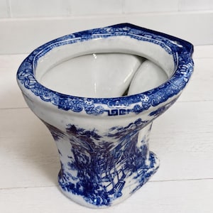 May include: A ceramic toilet bowl featuring a classic blue and white transferware pattern. The exterior and the rim are decorated with intricate landscape scenes and geometric borders, while the interior basin is finished in a plain white glaze.