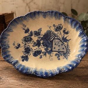 May include: An oval ceramic dish with a cream-colored base and a scalloped edge painted in deep blue. The center features a blue floral pattern with intricate botanical illustrations and a decorative fan-like element on a speckled off-white surface.