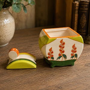 Peut inclure: Un récipient carré en céramique avec un couvercle amovible, présentant une base blanche, un bord orange et des accents verts. L'extérieur est orné de motifs floraux orange peints à la main et de feuilles vertes. Le couvercle a une poignée orange.