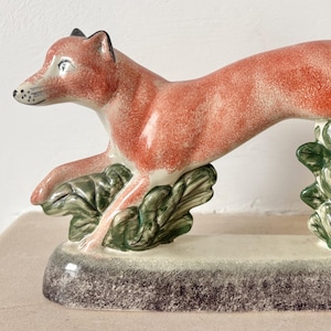 May include: A ceramic figurine of a leaping fox with a reddish-orange coat and black-tipped ears. The fox is positioned over two green leafy bushes, supported by a rectangular base with a dark, mottled gray finish and a smooth, glossy texture.