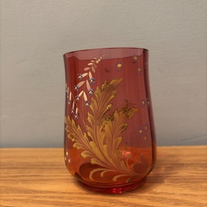May include: A ruby-coloured glass with a floral design. The glass features gold and white painted leaves and blue accents. A unique piece of glassware.