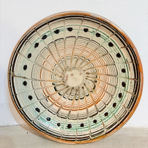 Könnte beinhalten: Ein dekorativer Keramikteller mit handgemaltem Design. Der Teller hat ein zentrales Spiralmuster in Weiß-, Orange-, Grün- und Schwarztönen mit schwarzen Punkten am Umfang. Der Rand ist in einem warmen Orangeton gehalten.