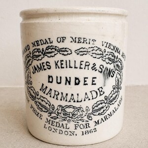 May include: A cylindrical off-white ceramic marmalade jar featuring black printed text that reads James Keiller & Sons Dundee Marmalade, along with Grand Medal of Merit Vienna 1873 and Prize Medal for Marmalade London 1862, surrounded by a decorative wreath.