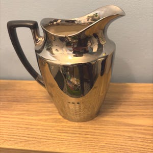 Puede incluir: Una cremera plateada con un asa negra. La cremera tiene una superficie reflectante y un pico curvo. El asa es de color oscuro y sólido. La cremera está sobre una superficie de madera.