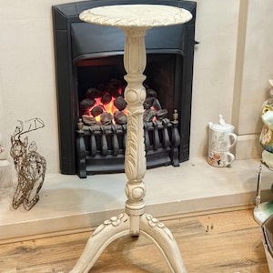 May include: Off-white, three-legged plant stand with a round top. The stand features carved floral and leaf details. The stand is placed in front of a fireplace with a black surround and a decorative metal grate.