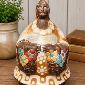 May include: A ceramic figurine of a person with a dark brown complexion, wearing a white shawl with a beaded collar. The figure holds two baskets filled with red, yellow, and turquoise flowers. The base features a white geometric pattern.