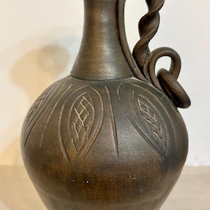 May include: A brown ceramic vase with a decorative handle and leaf-like patterns carved into the body. The handle has a twisted design. The vase has a dark, matte finish.