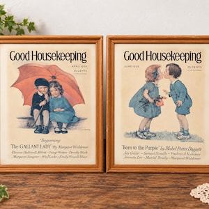 May include: Two framed vintage Good Housekeeping magazine covers from 1926. The left print shows children under a red umbrella. The right print depicts two children kissing. Both feature cream backgrounds, black text, and light brown wooden frames.