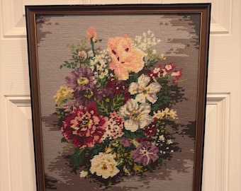 May include: Framed floral needlepoint art featuring a bouquet of colorful flowers. The flowers include shades of pink, red, white, yellow, and purple. The artwork is set against a gray background and framed in a brown wooden frame.