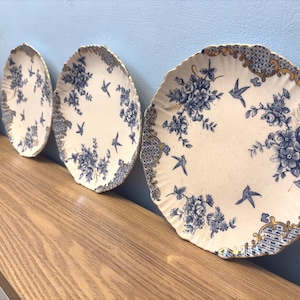 May include: Three antique decorative plates with a cream-colored background. Each plate features a blue floral and bird design. The plates are arranged on a wooden surface, with a blue wall in the background.