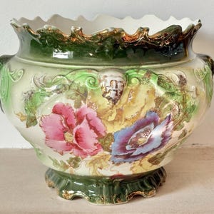Op de afbeelding: Een keramieken plantenbak met een geschulpte rand en voet, voorzien van een donkergroen glazuur en gouden accenten. De behuizing is crèmekleurig met reliëf bloemdetails, waaronder een grote roze bloem links en een blauwe bloem aan de rechterkant.