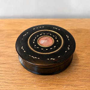 May include: A round, dark brown lidded box featuring a central circular inlay of polished pink stone. The lid is decorated with a thin gold-toned circular border and a repeating pattern of small, light-colored geometric inlays around the center.