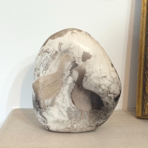 Può includere: Una scultura decorativa in pietra ovale con un motivo marmorizzato bianco e grigio. Due sagome di uccelli sono incise nella pietra. La scultura è esposta su una mensola di colore chiaro.