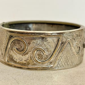 May include: Silver hinged bangle bracelet with detailed floral and geometric engraved patterns. The bracelet has a rectangular design and a decorative clasp. The metal has a slightly aged appearance.