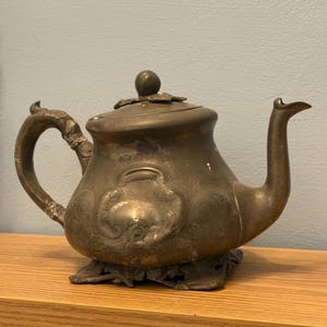 Puede incluir: Tetera antigua de peltre con detalles florales y de volutas. La tetera presenta un pico curvo, un asa decorativa y una tapa con un pequeño pomo. Se apoya sobre una base decorativa y se exhibe en una estantería de madera.