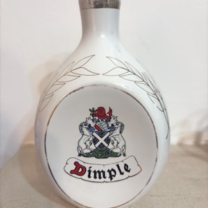 May include: White ceramic bottle with a gold-coloured top and a decorative crest. The bottle features a gold leaf design and the word "Dimple" in red script. A vintage-style liquor bottle.