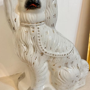 Op de afbeelding: Een witte keramische hondenfiguur met gouden accenten. De hond heeft een zwarte neus, een rode mond en gele ogen. Hij draagt een halsband met gouden details. Het beeldje zit rechtop en heeft grijze markeringen op zijn poten.