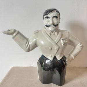 Könnte beinhalten: Keramikfigur eines Mannes in weißer Uniform mit schwarzem Schnurrbart und Monokel. Die Figur hat einen schwarzen Zylinder und eine Fliege. Der Mann posiert mit einem ausgestreckten Arm und dem anderen in der Hüfte.