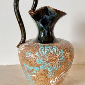 Puede incluir: Un jarrón decorativo de cerámica con un asa curva. El jarrón presenta una base dorada con diseños florales turquesa y blancos. La parte superior y el asa son de color marrón oscuro y el jarrón tiene una forma única.