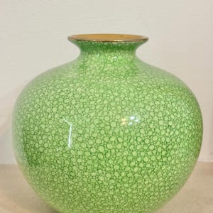 Pode incluir: Um vaso redondo de cerâmica verde com um padrão texturizado de pontos. O vaso tem uma borda dourada e um corpo largo e bulboso. O design é simples e elegante, adequado para exibir flores ou como peça decorativa. Altura aproximada 20 cm.