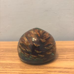 以下が含まれることがあります： 松ぼっくりが内包された、透明で丸みを帯びたペーパーウェイト。 松ぼっくりは茶色とタン色で、樹脂は基部に青緑色をしています。 ペーパーウェイトは木製の表面に置かれています。