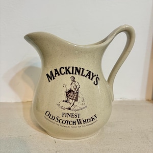 Op de afbeelding: Crèmekleurige keramische kan met een gebogen handvat en tuit. De kan heeft de tekst "MACKINLAY'S FINEST OLD SCOTCH WHISKY" met een illustratie van een man in een kilt. De kan heeft een gespikkelde afwerking.