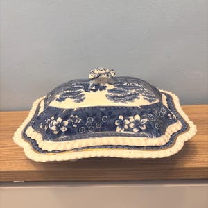 Może przedstawiać: Niebiesko-biała ceramiczna patera do serwowania z pokrywką. Naczynie ma wzór kwiatowy i krajobrazowy. Krawędzie są karbowane ze złotym wykończeniem. Pokrywka ma mały dekoracyjny uchwyt. Naczynie stoi na drewnianej powierzchni.