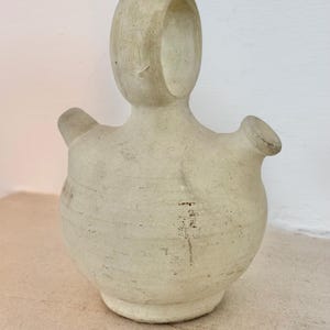 Puede incluir: Jarra de cerámica de color crema con un diseño único y abstracto. La jarra presenta un cuerpo redondeado, un asa alta de forma ovalada y dos picos. La superficie tiene un aspecto texturizado y envejecido, lo que sugiere una estética rústica o vintage.