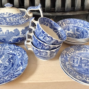 Puede incluir: Un juego de té de porcelana azul y blanca con tetera, tazas y platos. El juego tiene un diseño de paisaje. La tetera tiene tapa y boquilla curva. Los platos y tazas están apilados.