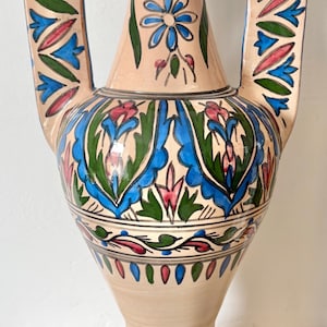 May include: A beige ceramic vase with two handles, decorated with blue, green, and red floral patterns. The vase has a blue rim and detailed designs around the body and base, displaying a traditional style.