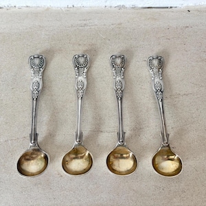 Op de afbeelding: Vier antieke zilveren lepels met sierlijke schelpvormige handvatten en goudkleurige kommen. De lepels zijn gerangschikt op een lichtgekleurd oppervlak, wat hun gedetailleerde ontwerp en vintage esthetiek benadrukt. Ideaal voor het serveren van specerijen of als decoratieve stukken.