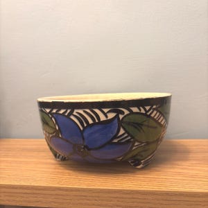May include: A small, ceramic bowl with a floral design. The bowl features a blue flower with green leaves and a black and white striped border. The bowl is supported by three small feet and sits on a wooden surface.
