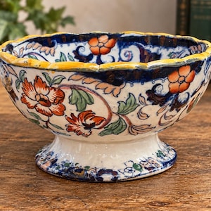 May include: A ceramic footed bowl with a scalloped rim, featuring a hand-painted floral pattern in orange, green, and dark blue on a white background. The bowl has a yellow painted edge and a wide, circular base with matching floral details.