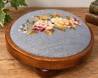 May include: A round wooden footstool featuring a blue fabric top with a needlepoint floral design. The stool has a dark brown polished wooden frame and short, rounded wooden feet, providing a low profile for this functional furniture piece.