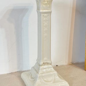 Peut inclure: Un bougeoir en céramique crème avec une base carrée et une haute colonne rectangulaire. La colonne présente des motifs floraux décoratifs et un sommet évasé. Le bougeoir mesure environ 25 cm de haut.