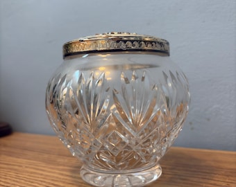 May include: A clear, cut-glass jar with a silver and gold-toned rim. The jar has a round body with a diamond pattern and sits on a small, clear base. The lid is silver and gold-toned. The jar is likely for storing small items.