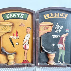 Peut inclure: Panneaux de toilettes en bois vintage, l'un indiquant "GENTS" et l'autre "LADIES". Le panneau "GENTS" représente un homme de dessin animé urinant, tandis que le panneau "LADIES" représente une femme de dessin animé. Les deux panneaux ont un aspect vieilli.
