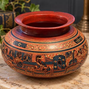 May include: A round, red lacquer bowl featuring intricate hand-painted details in black, gold, and blue. The exterior surface displays detailed scenes of figures within architectural settings, surrounded by decorative floral and geometric patterns.