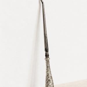 Puede incluir: Un ganchillo de plata antiguo con un extremo de gancho curvado y un eje largo y delgado. El mango presenta intrincados patrones florales y de volutas. Una herramienta de artesanía vintage.