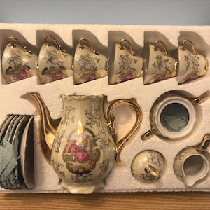 May include: A vintage tea set with a cream-colored finish and gold accents. The set includes a teapot, sugar bowl, creamer, six teacups, and saucers. Each piece features a decorative floral and figure design, presented in a protective foam insert.