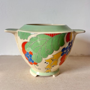 Puede incluir: Cuenco de cerámica color crema con borde y asas verdes. El cuenco está decorado con hojas verdes, detalles rojos y racimos de flores azules y amarillas. El cuenco tiene una base con pie.