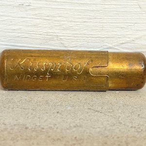 Puede incluir: Un estuche de lápiz labial dorado vintage con el texto "Kissproof Midget U.S.A." en relieve en el lateral. El estuche tiene forma rectangular con extremos redondeados y un mecanismo deslizante.