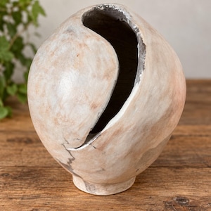 May include: A spherical vase with a smooth, light beige and white exterior finish. The vase features a prominent, curved vertical opening that reveals a dark, textured interior surface. The base is narrow and circular, supporting the rounded body.