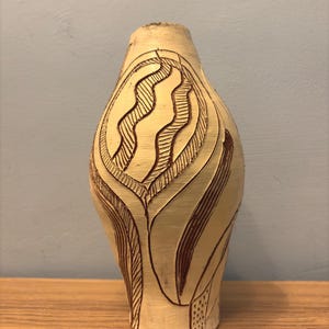 Op de afbeelding: Een beige keramische vaas met een gesneden ontwerp. De vaas heeft een bolvorm met een smalle hals. Het gesneden ontwerp heeft een reeks gebogen lijnen en bladvormige vormen. De vaas staat op een houten oppervlak.