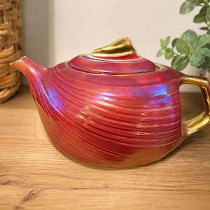 May include: A ceramic teapot featuring a vibrant red iridescent glaze with a textured, rippled surface. The teapot includes a gold-coloured handle and a matching gold-coloured finial on the lid, with a curved spout extending from the main body.