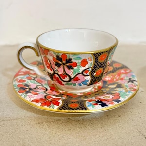 Op de afbeelding: Een decoratieve theekop en schotel set met een bloemenpatroon. De kop en schotel zijn versierd met rode, roze en blauwe bloemen, en afgewerkt met goud. De kop heeft een witte binnenkant en een gebogen oor. De schotel weerspiegelt het ontwerp van de kop.